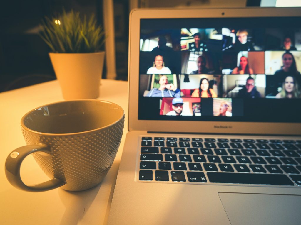 A photo of a laptop and coffee mug sitting on a table. The laptop screen shows a virtual meeting with small squares of virtual attendees.