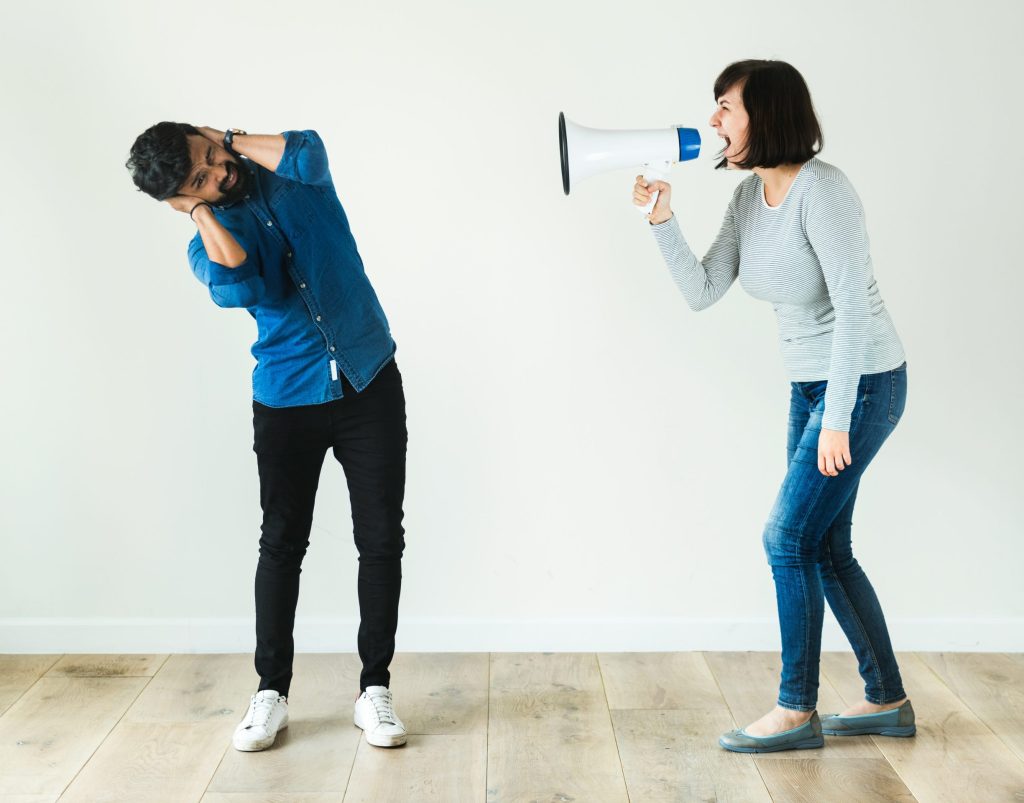 A woman holding a megaphone yells at a man standing next to her, as her covers his ears and leans away from her.